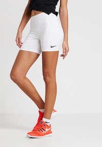 Pantalones cortos deportivos blancos con el logo negro de Nike, material elástico, diseño ajustado. Calzado naranja visible, realzando la apariencia deportiva.