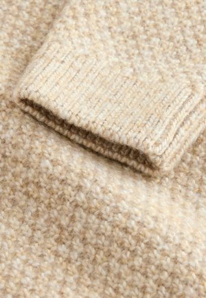 Beige Wollstoff mit einer strukturierten Oberfläche, die ein hell gesprenkeltes Muster aufweist. Der Bund zeigt ein geripptes Strickdetail, das die visuelle Tiefe verstärkt.