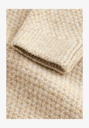 Tejido de lana beige con una superficie texturizada que presenta un patrón moteado claro. El puño muestra un detalle de punto acanalado, realzando su profundidad visual.