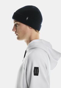 Young man in white hoodie and black knit beanie facing left with neutral expression on white background.
