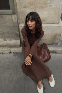 Robe marron à côtes avec un ourlet volé, associée à des ballerines blanches. Sac tissé marron posé à proximité. Modèle assis contre un mur texturé.