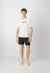 Biała bawełniana koszulka z logiem "CALVIN KLEIN" w kolorze czarnym, zestawiona z czarnymi szortami. Model stoi boso na tle jasnoszarym.