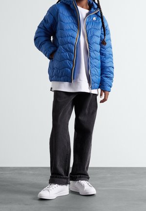 Person trägt eine blaue gesteppte Jacke über einem weißen Hoodie, schwarze Jeans und weiße Sneakers, steht auf einem grauen Boden vor einer hellen Wand.