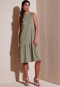 Sleeveless, knee-length dress in olive green ribbed fabric, featuring a tiered skirt and a rounded neckline. Worn with neutral strappy heels.
