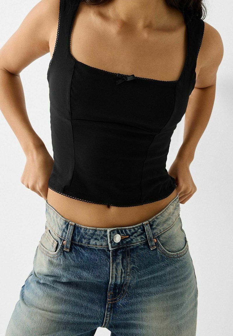 Femme portant un crop top noir ajusté sans manches associé à un jean bleu délavé taille haute sur un fond uni.