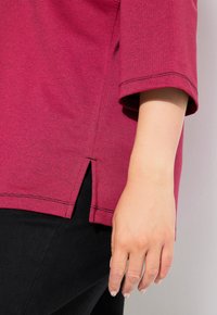 Top a righe bordeaux realizzato in tessuto leggero, con maniche corte e spacchi laterali all'orlo. Mano appoggiata su un indumento nero.