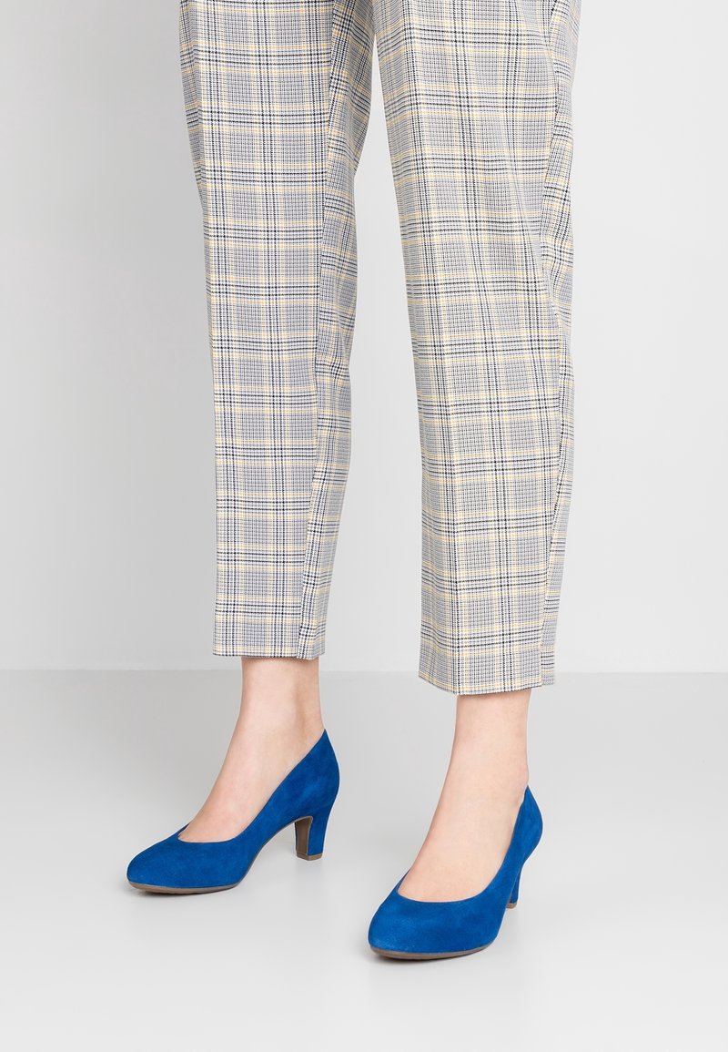 Des chaussures à talons hauts en daim bleu avec un bout rond et un talon bloc moyen, associées à un pantalon à motifs carreaux beige et marine.