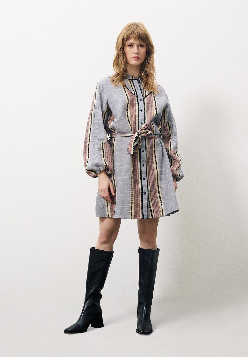 Robe grise rayée avec une taille ceinturée, un devant à boutons et des manches bouffantes. Des bottes noires montantes complètent le look.