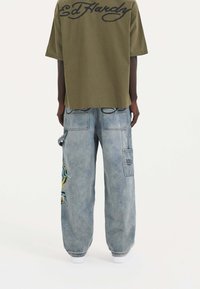 Olivgrön oversized t-shirt med svart logotyp på ryggen, matchad med ljusblå baggy jeans i denim med färgglad broderad design.