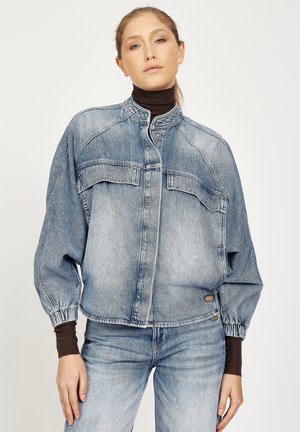 Woman wearing light blue oversized denim jacket with elastic cuffs over a brown turtleneck and matching light blue denim jeans.