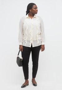 Γυναίκα με πλεξούδες που φοράει λευκό μπλουζάκι με βολάν, μαύρο παντελόνι, μαύρα χαμηλά τακούνια, κρατά μια μικρή μαύρη τσάντα, στέκεται και κοιτάζει προς το πλάι.