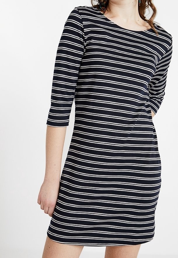 Kobieta w czarnej sukience w białe poziome paski i rękawami do trzech ćwierći, stojąca z jedną ręką w kieszeni na białym tle.