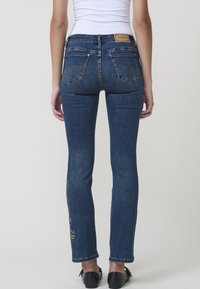 Modre denim hlače s ožjim krojem, ki imajo izvezene cvetlične motive na spodnjem delu hlačnic in klasičen dizajn s petimi žepi.