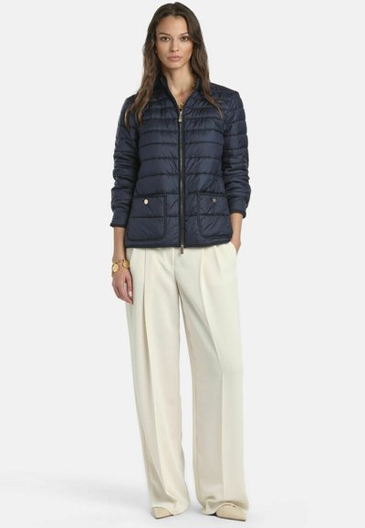 Donna con giacca trapuntata blu scuro con tasche frontali, pantaloni bianchi a vita alta e gamba larga, e scarpe beige con punta, in piedi con una mano in tasca.