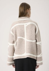Uldne beige sweater med hvide geometriske mønstre, brede ærmer og sjalkrave. Teksturen virker blød og lidt overdimensioneret.