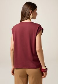 Haut maroon à manches courtes en coton doux avec des poignets roulés, associé à un pantalon beige. Boucles d'oreilles géométriques en or, au design simple.