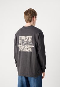 Tricou gri cu mânecă lungă, decorat cu un grafic mare, texturat în alb și negru, având inscripția "Barbour International England" pe spate.