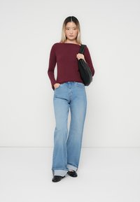 Bordeaux rood lang model shirt, lichtblauwe wijde jeans, en zwarte handtas. Model draagt zwarte ballerina's. Eenvoudige achtergrond, neutraal kleurenpalet.