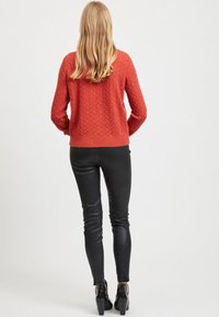 Kabelstrick orangefarbenem Pullover mit rundem Ausschnitt und gerippten Bündchen, kombiniert mit enganliegenden schwarzen Lederhosen und Absatzschuhen.