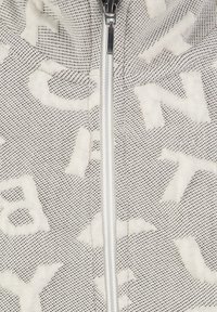 Primo piano di una zip bianca su tessuto grigio e bianco con texture e pattern di lettere astratte in rilievo.