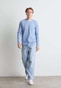 Camicia a maniche lunghe azzurro chiaro con scollatura rotonda, abbinata a jeans larghi azzurro chiaro e sneakers bianche, su uno sfondo semplice.