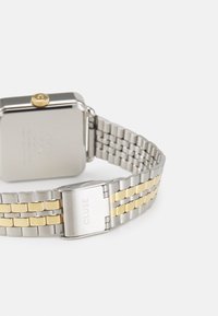 Montre en métal argenté et doré avec un cadran rectangulaire, un bracelet texturé et une couronne dorée. Elle possède un fermoir gravé "CLUSE".