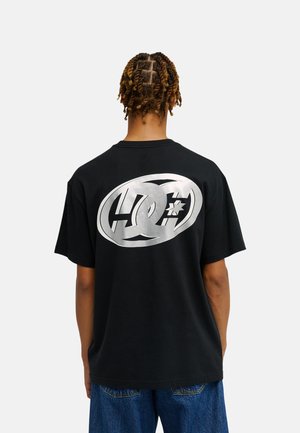 Osoba z poskręcanymi warkoczykami nosi czarny T-shirt z dużym srebrnym logo DC Shoes na plecach, do tego niebieskie dżinsy.
