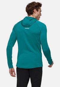 Mężczyzna odwrócony tyłem, ubrany w turkusową bluzę z kapturem i długim rękawem z logo "MAMMUT" na plecach, w połączeniu z czarnymi spodniami.