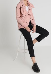 Giacca zip-up a pois rosa chiaro con colletto oversize sopra una maglietta bianca, abbinata a pantaloni neri cropped e sneakers nere.