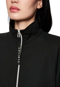 Veste zippée noire avec un col haut, un tissu texturé et un tirette zippée avec "SPORTS" imprimé en blanc sur une étiquette noire.