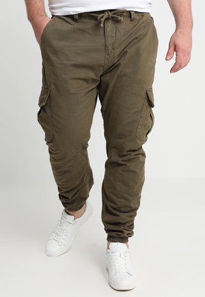 Mand iført olivengrønne cargobukser med elastiske manchetter og hvide sneakers, hænderne i lommerne, stående mod en ensfarvet baggrund.