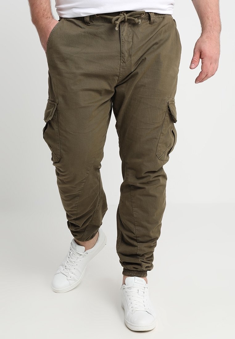 Homme portant un pantalon cargo vert olive avec des poignets élastiques et des baskets blanches, mains dans les poches, debout devant un fond uni.