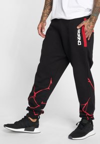 Black sweatpants with red abstract patterns on the sides, featuring an elastic waistband and cuffs, paired with black athletic shoes.