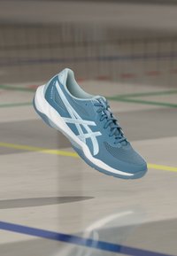 Μπλε και λευκό αθλητικό παπούτσι που αιωρείται πάνω από λαμπερό εσωτερικό δάπεδο αθλητικού γηπέδου με ορατές πολύχρωμες γραμμές γηπέδου.