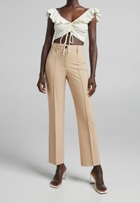 Krémový crop top s volánky a detaily na šňůrku, spárovaný s béžovými, vysokým pasem, rovnými kalhotami. Průhledné sandály na blokovém podpatku.