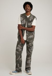 Chaleco de camuflaje y pantalones cargo en tonos verdes y grises, con una textura suave. Camiseta blanca y zapatillas azules completan el conjunto.