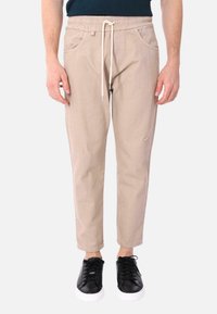 Pantaloni jogger beige con vita elasticizzata e cordoncino. Presentano tasche frontali e piccoli dettagli rovinati. Abbinati a sneakers nere.