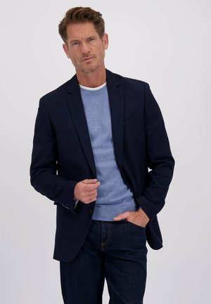 Marineblauw geblokt blazer over een lichtblauwe trui met een ronde hals. Donkere jeans maken de outfit af, wat een klassieke business casual stijl benadrukt.