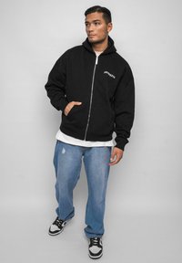 Hoodie noir à fermeture éclair en coton, avec une poche avant et un logo brodé. Associé à un jean clair déchiré et des baskets noires.