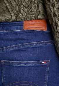 Mörkblå denimjeans med ribbad textur, med en brun läderetikett. Den stickade gröna tröjan har ett kabelmönster.