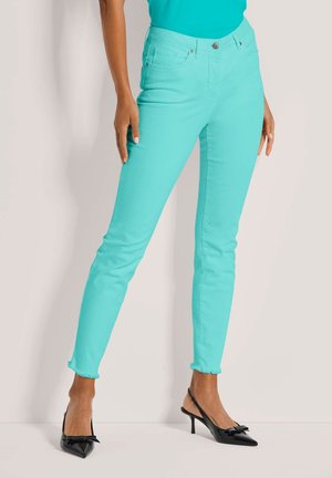 Vrouw draagt strakke aqua broek met rafelige zomen en zwarte puntige hakken met strikken, staand tegen een neutrale achtergrond.