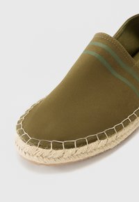 Olive green espadrille with a canvas upper, featuring two horizontal stripes. Jute rope midsole and stitched edges provide texture and detail.