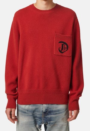 Homme portant un pull en tricot rouge avec un logo noir sur la poche poitrine gauche et un jean bleu clair déchiré.