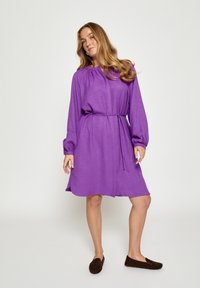 Lila Kleid mit langen Ärmeln, gerafftem Ausschnitt und Taillengürtel. Der Stoff hat eine strukturierte Oberfläche. Braune Slipper vervollständigen den Look.