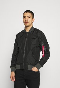 Svart bomberjacka i nylon med ribbade muddar och nederkant. Har en framdragkedja, sidofickor och en avtagbar röd etikett på ärmen.