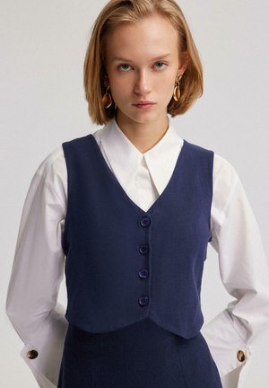 Gilet en mélange de laine bleu marine avec un décolleté en V, comportant quatre boutons ronds violets et un design court, porté sur une chemise blanche à col.