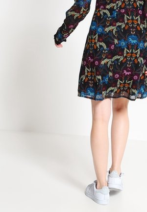 Femme portant une robe noire à fleurs et des baskets blanches, debout sur un fond clair uni, vue de la moitié du dos vers le bas.