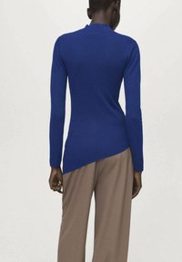 Rebrasti modri vrh z dolgimi rokavi in asimetričnim robom. V kombinaciji z bež hlačami. Poudarek na teksturi in živahnem kontrastu barv.