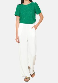 Grüner Spitzenoberteil mit floralem Ausschnitt, kombiniert mit lockeren weißen Schlaghosen. Die Sandalen haben einen mehrfarbigen, gemusterten Riemen.