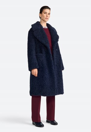 Cappotto teddy blu navy, morbido e peloso, con grandi revers, due tasche frontali e bottoni doppi. Indossato su un completo bordeaux.